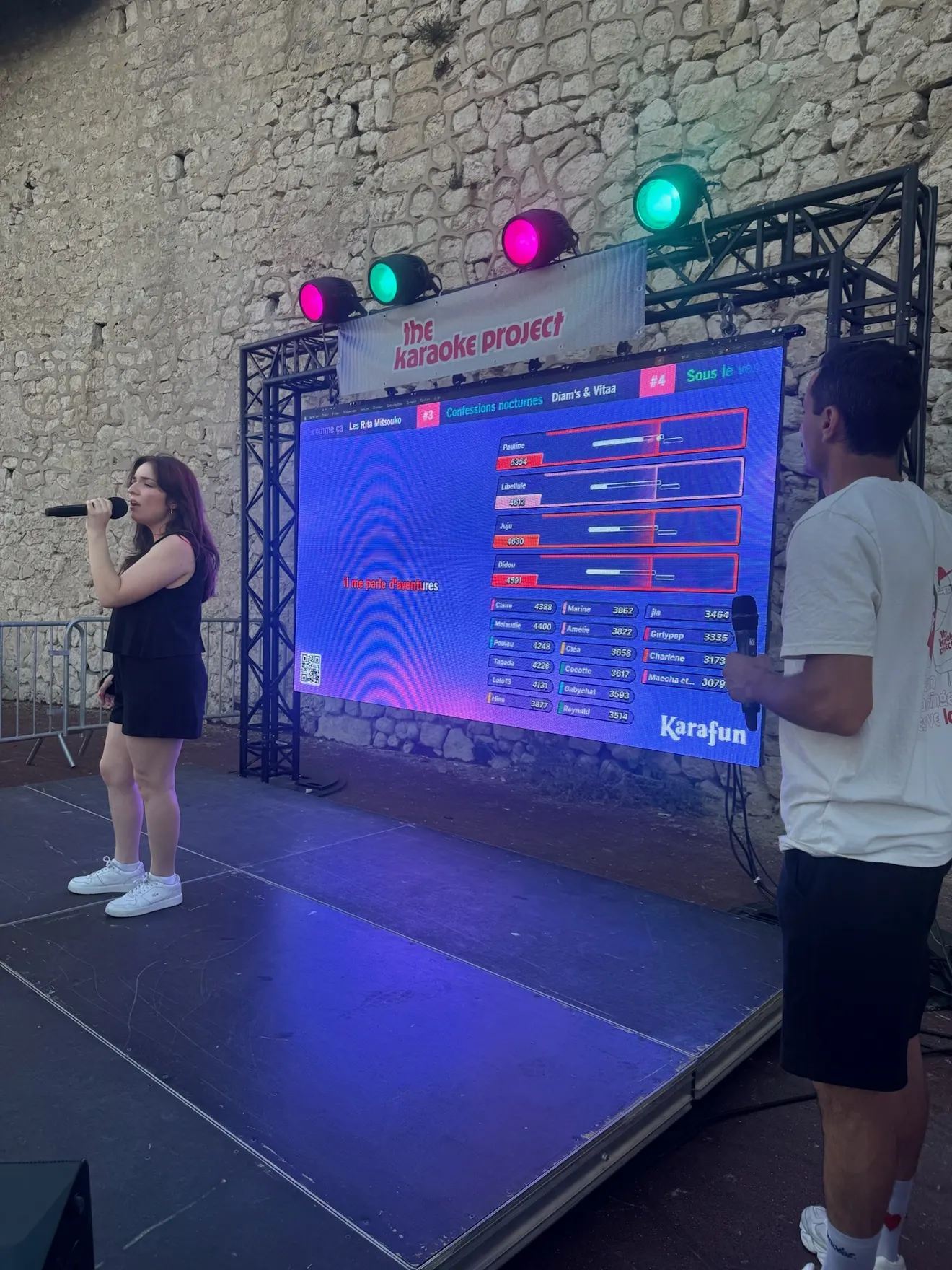 Une candidate chante sur scène, l’animateur à côté, les scores en direct sur l’écran